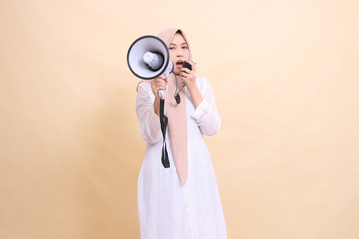 Asian Muslim woman wearing a hijab shouts at the camera sullenly, holding a loudspeaker megaphone and holding the speaker mic near the center's mouth. Technology, broadcast and promotion concept