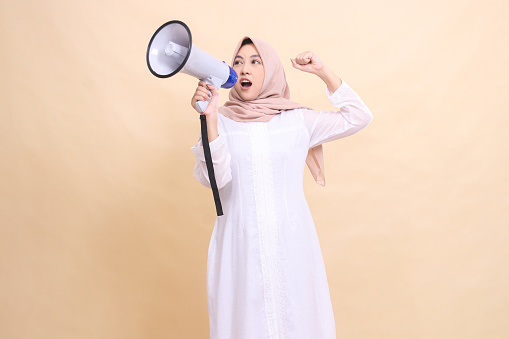 girl indonesia wearing hijab slanted to the right candidly shouts enthusiastically, holding a loudspeaker megaphone and clenching her fists upwards. Technology, broadcast and promotion concept