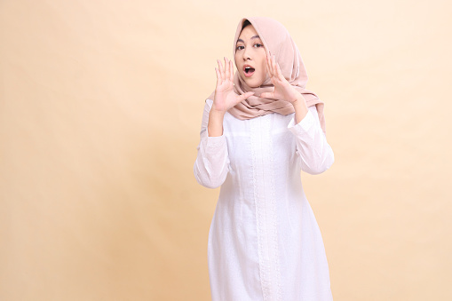 woman asian muslim standing to the camera with both hands near her mouth shouting an important announcement. Fashion, pointer, business and promotion concept