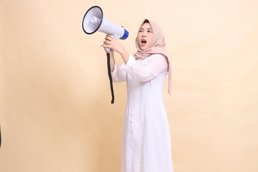 woman asian mature wearing hijab slanted to the right candidly shouting loudly, holding a loudspeaker megaphone with both hands. Technology, broadcast and promotion concept