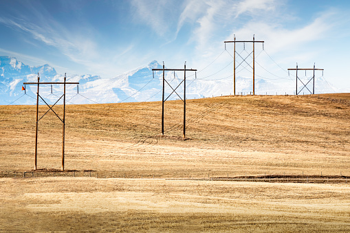 Power lines hanging from large wooden transmission towers over a hilltop overlooking distant mountain tops of the Canadian Rockies in Rocky View County Alberta Canada.