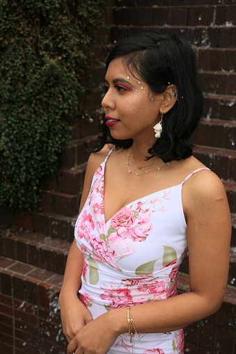 A closeup of a Bangladeshi woman standing in front of a manmade water feature. She is wearing medium length black hair, makeup, decoration, earring, jewelry and a white, spaghetti strap print dress.
