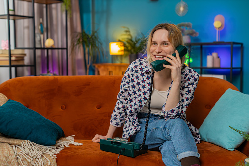Smiling young woman making wired telephone call conversation with friends sitting on couch at home in room. Happy excited pretty girl enjoying old-fashioned retro phone from 90s talking in living room