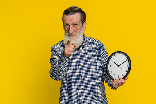 It is your time, hurry up. Senior old man showing time on wall office clock, ok, thumb up approve pointing finger at camera, advertisement, deadline. Mature grandfather isolated on yellow background