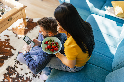 Mid adult interracial couple sitting in their living room, looking at mobile phone and eating strawberries. They are having relaxing time, looking happy.