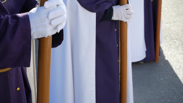 Closeup of three penitent in line formation dressed on traditional white and purple tunics ready to march. They hold long candles with gloves. Camera tilts down showing their robe's. Holy week.