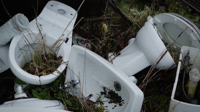 Abandoned white toilets with plants growing though them. Handheld detail of rubbish