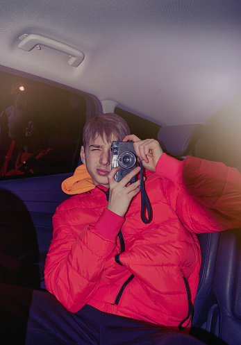 Young teen in red puffer jacket sits in a car, taking a picture with film camera. The 90s scene frontal photo.