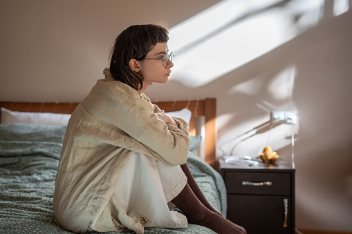 Sad tired teen girl sits on bed at home looking at window feeling apathy. Blue mood of depressed girl. Nervous female in frustration hiding from society in loneliness. solitude, life problems troubles