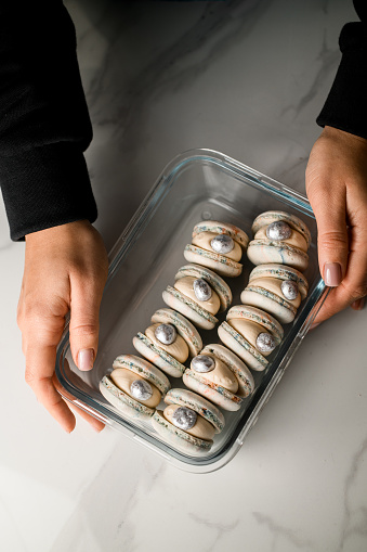 Female hands holding a rectangular glass container with beautiful shell-like macaroons standing on a white kitchen table