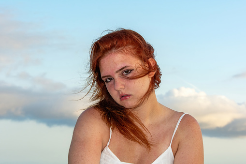 Portrait of a young girl with red hair, blue sky and clouds, hair tousled by the wind