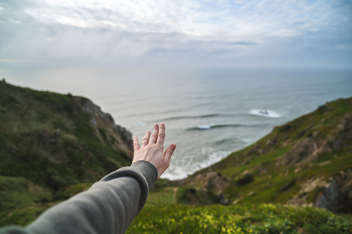 Woman's hand reaches out towards the ocean from a cliff. Human connection with nature concept. Shot near Cabo da Roca, Portugal.