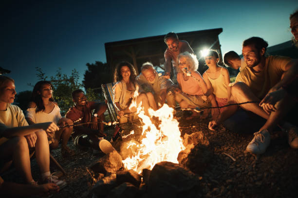 Group of mixed age group of people relaxing by a bonfire and cooking marshmallow candy
