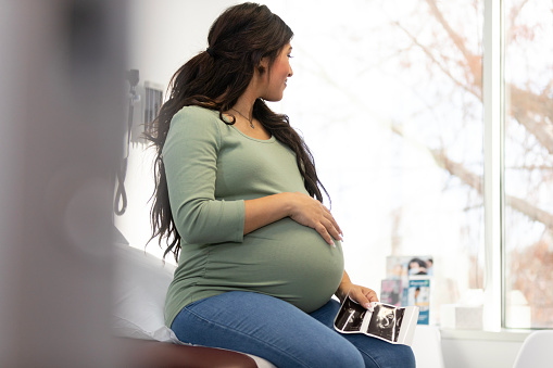 The young adult pregnant woman holds the ultrasound photos of her unborn baby as she looks out the window and daydreams.