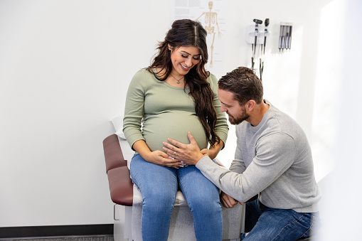 As she sits on the examination table, the young adult woman looks down and smiles at her young adult husband trying to feel the baby moving.