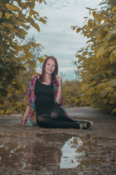 A young woman with red hair is biting sunglasses and posing while sitting down on a path full of leaves around her. Woman surrounded by lush greens and water reflection. Sexy modern outdoor woman