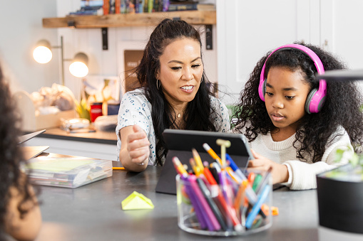 While an unrecognizable daughter seated across the table works independently, the mid adult mother helps her elementary age daughter with a mobile app on the digital tablet.