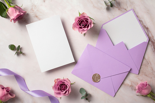 Wedding invitation card mockup, violet envelopes, rose flowers, eucalyptus leaves on marble table. Flat lay, top view, copy space.