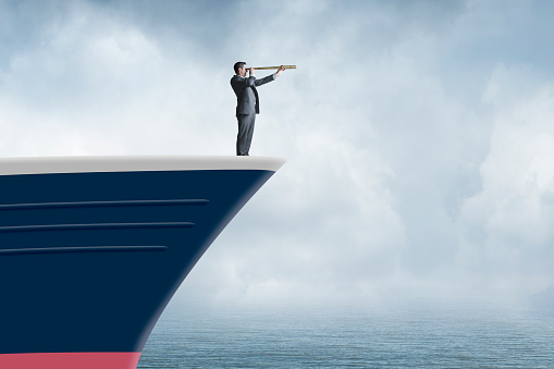 A businessman standing at the bow of a ship looks through a spyglass into the distance.