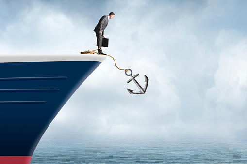 A businessman standing at the bow of a ship while holding his briefcase looks over the edge as an anchor drops into the water while it is attatched to his leg.