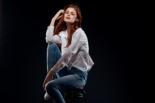 pretty woman sitting on a chair posing white shirt jeans long hair red lips dark background. High quality photo