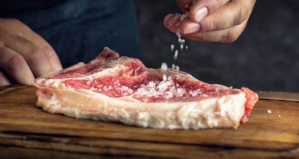 Homem polvilha sal em um bife suculento em uma tábua de corte de madeira Homem polvilha sal em um bife suculento em uma tábua de corte de madeira - fotos, fotos e imagens de carne crua