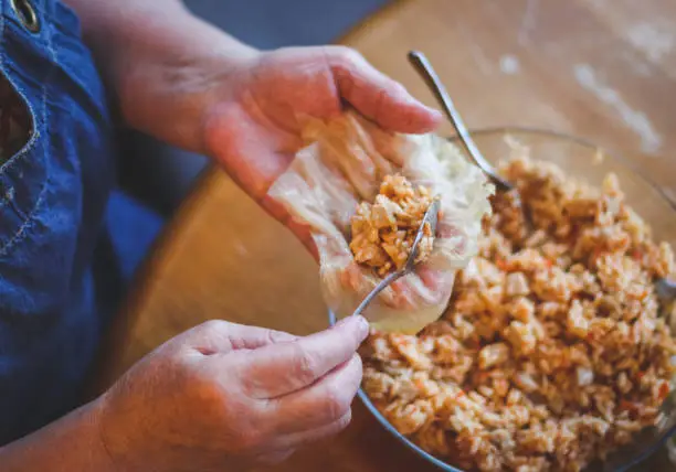 The hands of a senior woman hold a raw leaf of sauerkraut and put a metal teaspoon of rice-meat stuffing, sitting at a round table in the kitchen, top view close-up. The concept of step by step instructions, home cooking, traditional recipes, national cuisine, cabbage rolls, dolma.