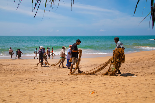 Unawatuna, Sri Lanka 09 february 2023 group of men in summer clothes of different colors helping fishermen to pull over large fishing net from ocean at beach of south Sri Lanka. hard team work.