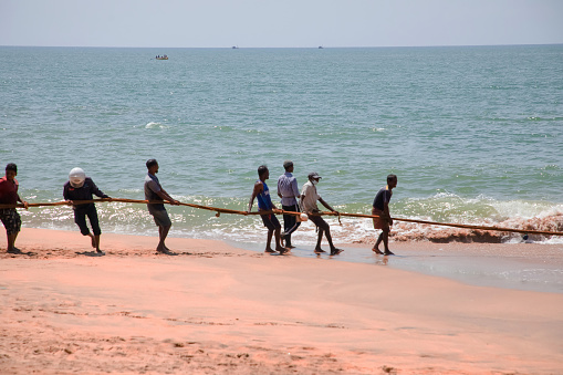 Unawatuna, Sri Lanka 09 february 2023 group of men in summer clothes of different colors helping fishermen to pull over large fishing net from ocean at beach of south Sri Lanka. hard team work.