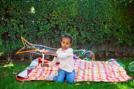 Happy African-American girl with black curly hair blowing and playing with soap bubbles at green garden