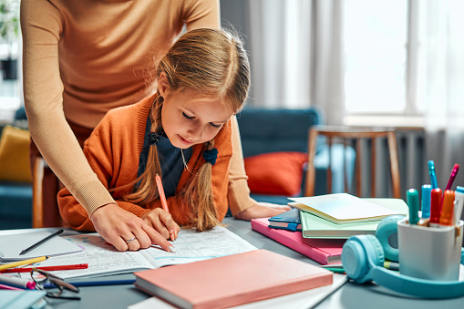 Time of homework checking. Unrecognizable person pointing with finger at schoolgirl's diary on desk with stationery. Blonde girl filling in tables with colourful pen while sitting at home.