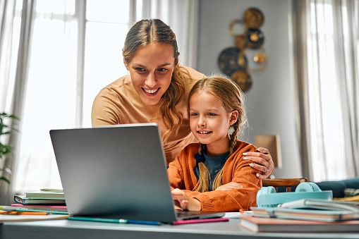Online training for children. Interested parent looking over daughter's shoulder into portable computer screen. Experienced child setting application for connecting to distant lesson from home.