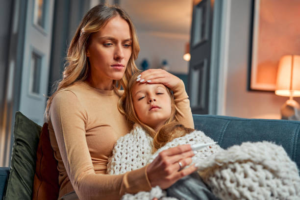 Mother caring about kid. Mom measuring ill daughter's temperature and looking at thermometer while sitting on couch Mother caring about kid. Frightened mom measuring ill daughter's temperature and looking at thermometer while sitting beside girl. Parent taking care of unhealthy child sleeping on couch. fever stock pictures, royalty-free photos & images