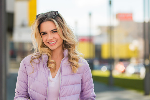 Against the backdrop of a bustling modern cityscape, a confident young woman with beautiful curly blonde hair looks directly at the camera, exuding charisma and poise. Her captivating smile and confident demeanor add a touch of urban sophistication to the scene, making this image ideal for conveying confidence, modernity, and urban allure.