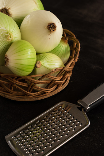 Some big green onions to cook in a brown basket and a metal grater on a black table with copy space