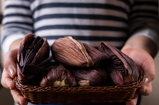 Woman holding many latin traditional food made of corn and wrapped in purple corn leaves in a basket on a black wooden table
