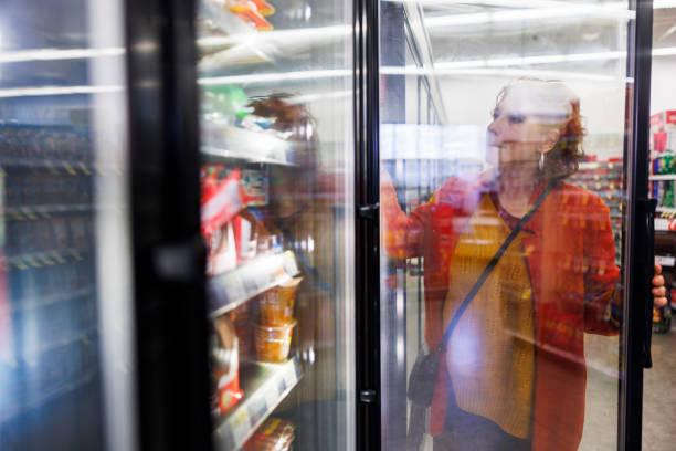 Mature Caucasian redhead woman selecting products from frozen food aisle in supermarket. Woman opening the glass door and picking the product. Looking through the glass