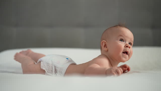 Side view of sweet infant baby wearing white diaper lying on stomach and happily waves arms and legs with smile. Portrait of nice newborn child learning to hold head