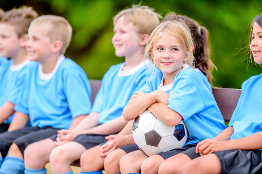 A small group f school-aged children sit on a bench along the side of a soccer field as they wait for the game to start.  They are each wearing jerseys and one child is holding the soccer ball.