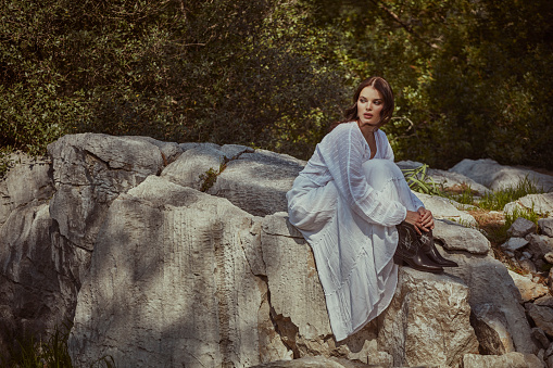 A woman in a white dress sitting in nature, among the trees, on the rocks.
