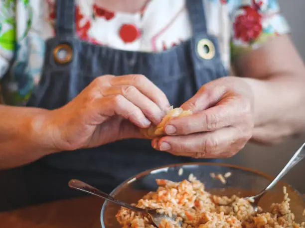 Hands of a senior woman wrapping a leaf of sauerkraut with rice-meat filling, sitting at a round table in the kitchen, close-up side view. The concept of step by step instructions, home cooking, traditional recipes, national cuisine, cabbage rolls, dolma.