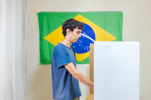 In this image depicting Brazilian elections, a young Brazilian man is seen casting his vote on an electronic ballot box in a voting booth. A Brazilian flag hangs on the wall of the polling place