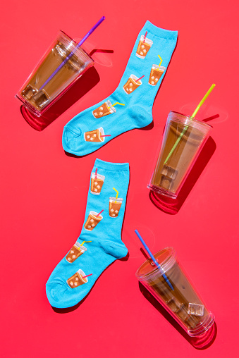 Flay lay of a pair of socks with an iced coffee pattern, surrounded by cups of iced coffee on a bright magenta background
