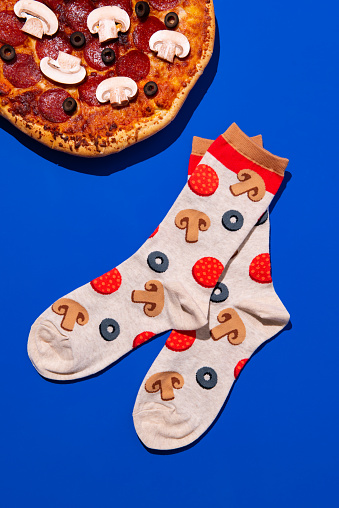 Flay lay of a pair of socks with a pizza topping pattern next to a pizza pie with those same toppings on a vibrant blue background