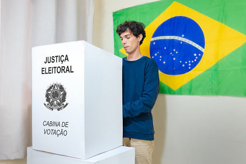 In this image depicting Brazilian elections, a young Brazilian man is seen casting his vote on an electronic ballot box in a voting booth. A Brazilian flag hangs on the wall of the polling place