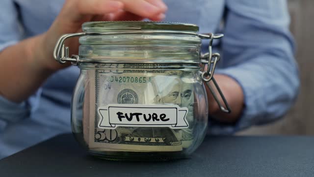 Female hands Saving Money turn Glass Jar filled with Dollars banknotes. FUTURE transcription in front of jar. Managing personal finances extra income for future insecurity