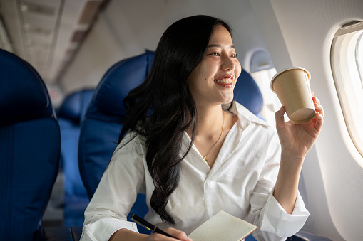 An attractive, elegant Asian businesswoman is sipping coffee while looking at the view outside the plane window, traveling on a business trip. businesspeople, transportation, luxurious lifestyle