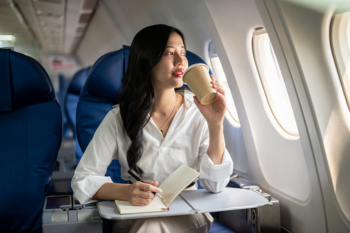 An attractive, elegant Asian businesswoman is sipping coffee while looking at the view outside the plane window, traveling on a business trip. businesspeople, transportation, luxurious lifestyle