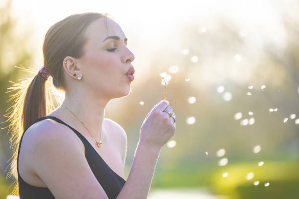 A woman blowing on a dandelion and making a wish outdoors.