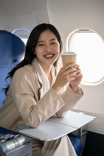 An attractive Asian businesswoman in a business suit is smiling at the camera, sitting at a window seat with her coffee cup, traveling by plane for a business trip.